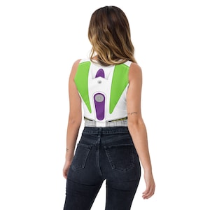 May include: White crop top featuring a Buzz Lightyear costume design. The top has green and purple accents, with a silver-colored circular detail in the center. The design mimics the character's space suit, with a printed belt detail at the bottom.