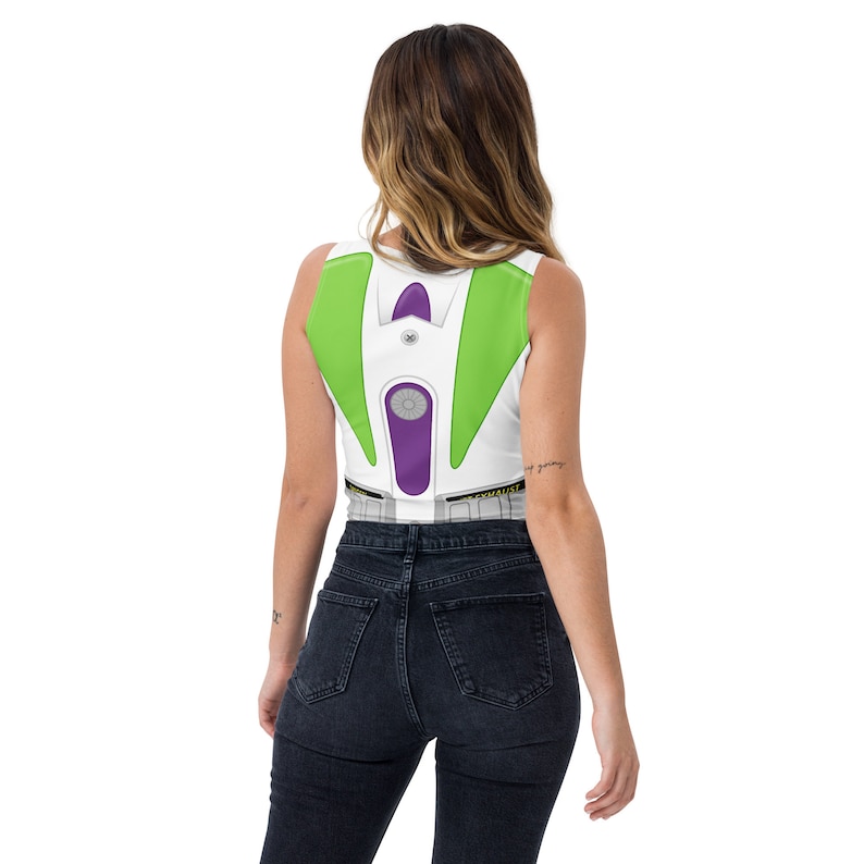 May include: White crop top featuring a Buzz Lightyear costume design. The top has green and purple accents, with a silver-colored circular detail in the center. The design mimics the character's space suit, with a printed belt detail at the bottom.