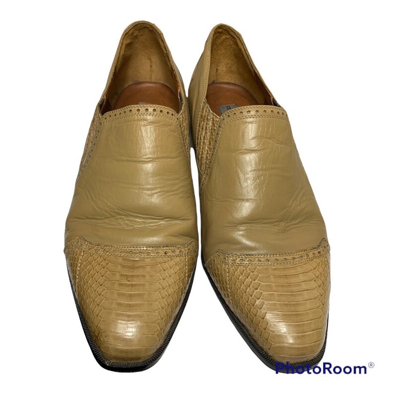 May include: A pair of tan leather slip-on shoes. The shoes have a textured, reptile-like pattern on the toe and side panels. The vamp and heel are smooth leather. The shoes have a pointed toe and a low heel.