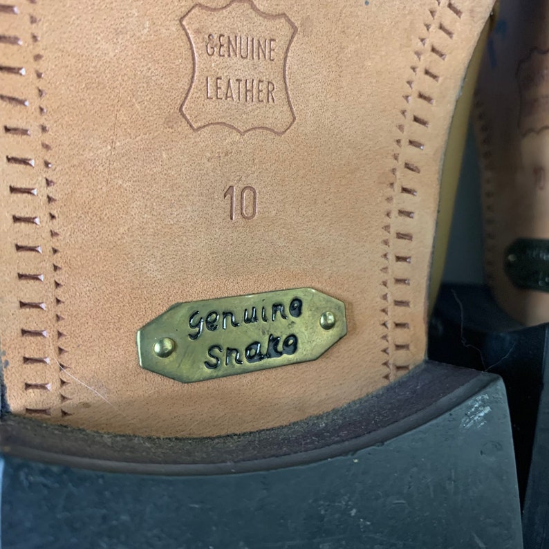 May include: Close-up of the sole of a shoe. The sole is tan leather with the words "GENUINE LEATHER" embossed. A metal tag reads "Genuine Snake". The number "10" is stamped on the leather.