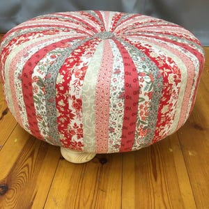 May include: A round, multi-coloured fabric ottoman with a tufted top. The ottoman is made of a patchwork of floral fabric in shades of red, pink, and grey. The ottoman has a wooden base.