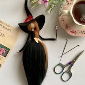 May include: A needle felted witch doll with long black hair, a black dress, and a black witch hat with a red flower. The doll is on a white surface with felting needles, scissors, and a cup of tea.