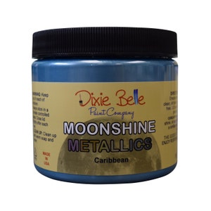 Pode incluir: Um frasco de vidro de tinta Moonshine Metallics da Dixie Belle na cor Caribe. O frasco tem uma tampa preta e um r&oacute;tulo amarelo com o logotipo da Dixie Belle e as palavras "Moonshine Metallics" e "Caribe".