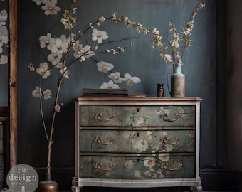 Puede incluir: Una cómoda de tres cajones con un acabado blanco desgastado y un patrón floral. La cómoda tiene herrajes dorados y se encuentra sobre un suelo gris. El fondo es una pared gris azulada con un diseño de papel tapiz floral.