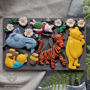 May include: A gray silicone mold featuring Winnie the Pooh characters, including Winnie the Pooh, Piglet, Tigger, and Eeyore. The mold also includes daisies, bees, and a honey pot. The text "re-design with prima" and "#681876 Decor Moulds Olde Bear Lane" are visible.