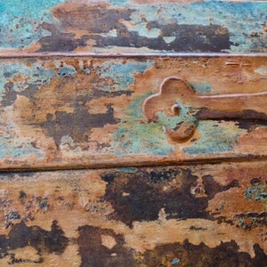 May include: Close-up of a wooden surface with a distressed, weathered look. The surface is painted in shades of blue, green, brown, and rust. A metal hinge is attached to the surface.
