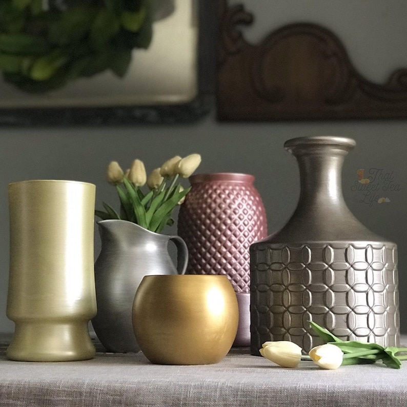 Pode incluir: Cinco vasos decorativos em diferentes cores e estilos, incluindo dourado, prateado e marrom. Os vasos est&atilde;o dispostos em uma mesa com uma toalha de mesa marrom claro. Os vasos s&atilde;o todos de diferentes formas e tamanhos, e todos s&atilde;o decorados com padr&otilde;es diferentes.