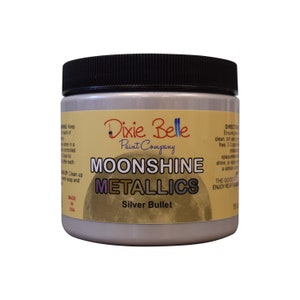 Pode incluir: Um frasco de tinta met&aacute;lica prateada com um r&oacute;tulo amarelo que diz "Dixie Belle Paint Company Moonshine Metallics Silver Bullet".