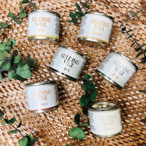 May include: Six small metal containers of gilding wax in various colors, including gold, copper, black, zinc, bronze, and silver. The containers are arranged on a brown paper background with green leaves.