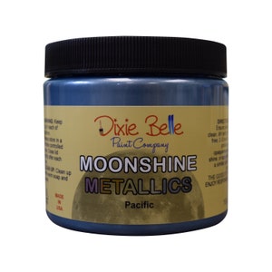Pode incluir: Um frasco de tinta met&aacute;lica Moonshine da Dixie Belle na cor Pacific. O frasco &eacute; azul com um r&oacute;tulo dourado que diz "Dixie Belle Paint Company" e "Moonshine Metallics".