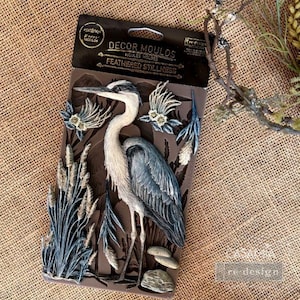 May include: A brown silicone mold featuring a detailed heron design with the text "FEATHERED STILLNESS". The heron is depicted in shades of blue, gray, and white, surrounded by reeds and floral accents. The mold is designed for crafting and decorative purposes.