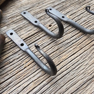 May include: Three hand-forged metal hooks with a rustic, industrial aesthetic. Each hook has a flat mounting plate with two holes and a curved hook. The hooks are a dark gray color and appear to be made of iron. The hooks are displayed on a weathered wooden surface.