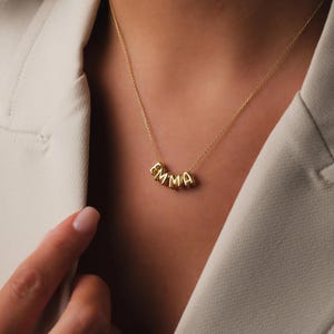 Könnte beinhalten: Eine goldene Halskette mit dem Namen "EMMA", der in einzelnen Buchstaben geschrieben ist. Die Buchstaben sind durch eine zarte Kette verbunden.