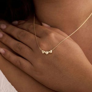 May include: A gold necklace with three heart pendants, each bearing an initial. The necklace is shown against skin, emphasising the design and colour. The chain is fine, and the hearts are evenly spaced.