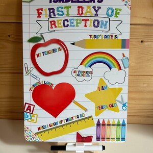 May include: A rectangular white dry-erase board with a lined paper background featuring colourful school-themed graphics including an apple, a rainbow, a star, and crayons. The text reads Isabella's First Day of Reception with fillable sections.
