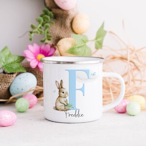 Op de afbeelding: Witte emaille mok met een blauwe letter F en een konijn dat een pot bloemen vasthoudt. De mok heeft de naam Freddie onder de letter gedrukt.