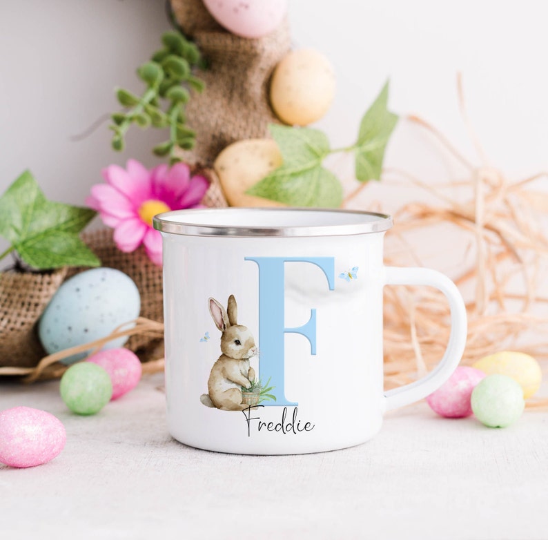 Op de afbeelding: Witte emaille mok met een blauwe letter F en een konijn dat een pot bloemen vasthoudt. De mok heeft de naam Freddie onder de letter gedrukt.