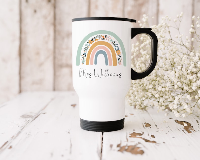May include: White travel mug with a black lid and handle. The mug features a rainbow with floral accents and the text "Mrs. Williams".