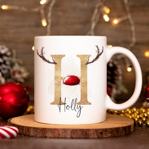 May include: White ceramic mug with a gold glitter letter "H" with reindeer antlers and a red nose. The mug has the name "Holly" written below the letter.