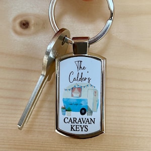 May include: A silver keyring with a white rectangular tag featuring a cartoon illustration of a blue and white caravan with a red awning. The tag also includes the text "The Calders Caravan Keys".