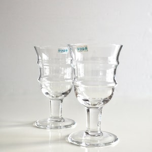 May include: Two clear, stemmed glasses with a unique, ridged design on the bowl. The glasses have a wide, circular base and a slender stem. Each glass has a small, rectangular sticker near the top. The background is plain white.