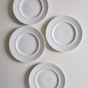 May include: Four circular white ceramic plates arranged in a cluster. Each plate features a smooth, flat centre basin surrounded by a wide, raised rim decorated with a repeating pattern of vertical, narrow, parallel ridges along the entire edge.