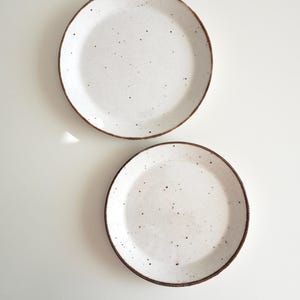 May include: Two white ceramic plates with a speckled glaze and brown rim. The plates are on a white background.