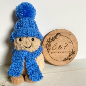 May include: A small, beige plush doll with a smiling face, wearing a bright blue crocheted hat with a pom-pom and a matching blue scarf. Beside the doll is a round wooden disc engraved with a leaf wreath and the text E & F Edward and Fluff.