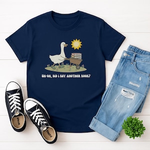 Può includere: T-shirt blu navy con un'oca dei cartoni animati che spinge una carriola piena di libri sotto un sole giallo. Il testo "Uh-oh, did I buy another book?" è stampato sotto. Sono visibili anche scarpe da ginnastica nere e jeans blu.