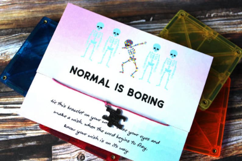 May include: A white card with the text "NORMAL IS BORING" and a pink string bracelet with a silver puzzle piece charm. The card also has a handwritten message about making a wish when the cord begins to fray.