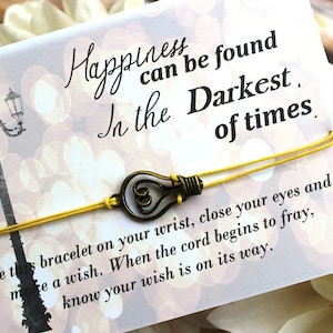 May include: A yellow string bracelet with a light bulb charm. The bracelet is tied around a card with the text "Happiness can be found in the Darkest of times. Tie this bracelet on your wrist, close your eyes and make a wish. When the cord begins to fray, know your wish is on its way."