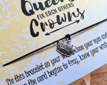 Peut inclure: Un bracelet en cordon noir avec un charm en forme de couronne argentée. La carte derrière le bracelet porte l'inscription "Queens fix each others crowns". Le fond est jaune pâle avec des mouchetures blanches.