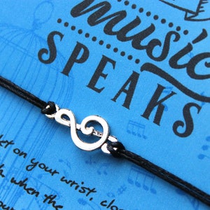 Peut inclure: Un bracelet en cordon noir avec un charme de cl&eacute; de sol argent&eacute;. Le bracelet est sur un fond bleu avec le texte "music speaks" en lettres noires.