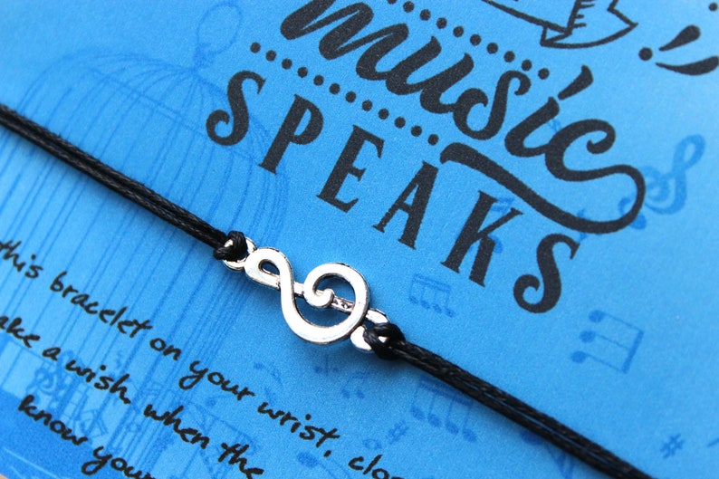 Peut inclure: Un bracelet en cordon noir avec un charme de cl&eacute; de sol argent&eacute;. Le bracelet est sur un fond bleu avec le texte "music speaks" en lettres noires.