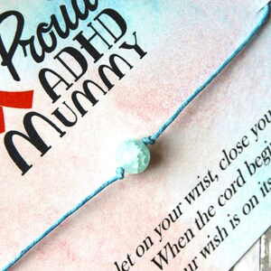 Puede incluir: Un brazalete de cuerda azul con una cuenta azul claro y blanca. El brazalete est&aacute; en una tarjeta con el texto "Proud ADHD Mummy" y una cinta roja.