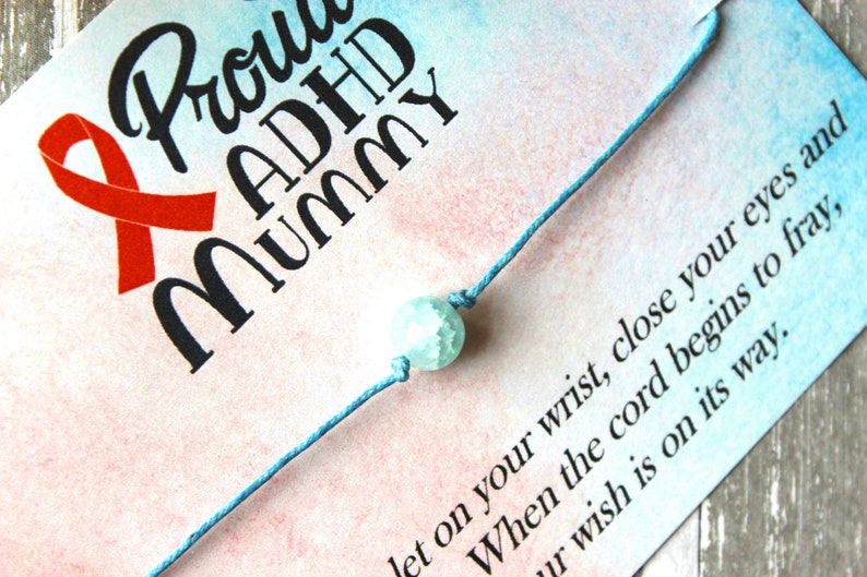 Puede incluir: Un brazalete de cuerda azul con una cuenta azul claro y blanca. El brazalete est&aacute; en una tarjeta con el texto "Proud ADHD Mummy" y una cinta roja.