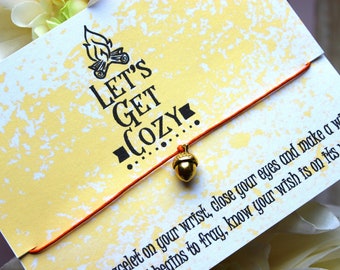 Peut inclure: Un bracelet en cordon rouge avec un charme d'écureuil en or et une carte qui dit "Let's Get Cozy." La carte a un fond jaune avec un motif de peinture blanche.
