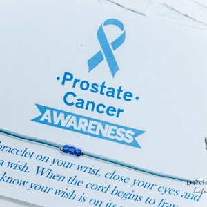May include: A blue ribbon tied to a blue string bracelet with a blue bead. The card reads "Prostate Cancer AWARENESS" and "Tie this bracelet on your wrist, close your eyes and make a wish. When the cord begins to fray know your wish is on its way." The card also has a small lavender flower graphic and the text "Dalvin's Baubles handmade with love."