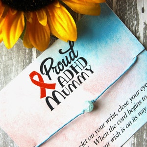 Puede incluir: Un brazalete de cuerda azul con una cuenta turquesa y una tarjeta que dice "Proud ADHD Mummy" con una cinta roja. La tarjeta tiene un fondo de acuarela rosa y azul.