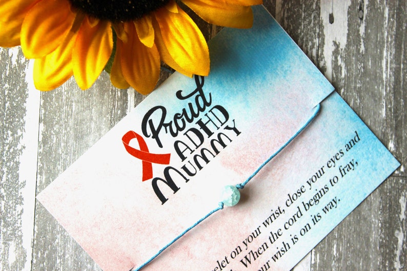 Puede incluir: Un brazalete de cuerda azul con una cuenta turquesa y una tarjeta que dice "Proud ADHD Mummy" con una cinta roja. La tarjeta tiene un fondo de acuarela rosa y azul.