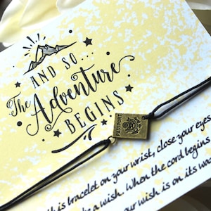 May include: A yellow card with black text that reads "AND SO... The Adventure Begins" with a small mountain illustration and stars. A black cord bracelet with a gold charm that says "PASSPORT" is attached to the card. The card also has text that reads "This bracelet on your wrist, close your eyes and make a wish. When the cord begins our wish is on its way."