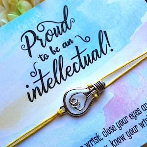 May include: A yellow string bracelet with a silver lightbulb charm. The bracelet is tied around a card with a watercolor background and the text "Proud to be an intellectual!"