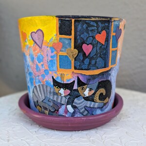 May include: A hand-painted ceramic flower pot with a matching purple saucer. The pot features a vibrant scene of two stylized cats resting near a window, surrounded by colorful floating hearts against a yellow, blue, and purple background.