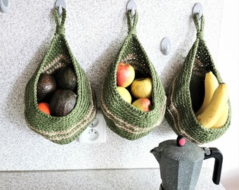 May include: Three teardrop-shaped hanging baskets made of green and tan woven cord, each suspended from a wall hook. The baskets contain avocados, oranges, apples, and bananas. A grey speckled moka pot sits on the counter below the baskets.