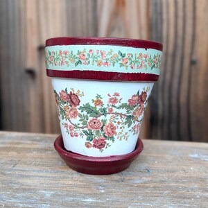 May include: A white ceramic flower pot with a matching saucer, featuring a dark red rim and base. The pot is decorated with a floral pattern of pink and red roses with green leaves, and a smaller floral border around the top rim.