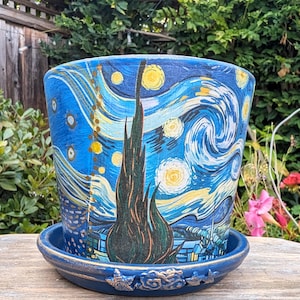 May include: A cylindrical ceramic plant pot and matching saucer featuring a hand-painted reproduction of a starry night sky with swirling blue clouds, bright yellow stars, and a prominent dark green cypress tree silhouette in the center.