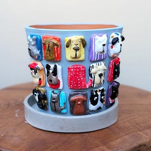 May include: A light blue cylindrical planter featuring a collection of small, colorful, three-dimensional dog faces attached to the exterior. The planter sits on a matching light blue circular saucer, showcasing various breeds and patterns in relief.