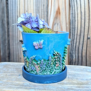May include: A light blue ceramic plant pot with a dark blue saucer, featuring a raised relief design of green foliage, colorful wildflowers, and a small butterfly on the exterior, containing a cluster of purple hydrangea flowers.