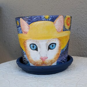 May include: A cylindrical ceramic plant pot with a matching dark blue saucer. The exterior features a hand-painted orange and white cat wearing a yellow hat, set against a dark blue background with swirling yellow stars and circular patterns.