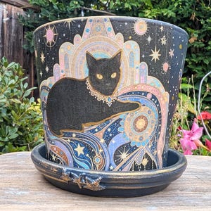 May include: A black ceramic plant pot with a matching saucer, featuring a detailed illustration of a black cat wearing a jeweled collar. The design includes celestial motifs, stars, and intricate patterns in shades of blue, pink, and gold.